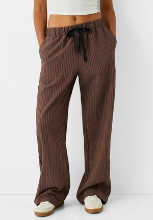 Personne portant un pantalon marron ample avec de fines rayures verticales noires, une ceinture noire à la taille, un crop top blanc et des baskets blanches avec semelles en gomme.