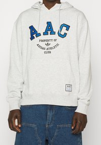 Grå huvtröja med dragsnöre i huven. Har "AAC" i blå och svart text, samt "ADIDAS ATHLETIC CLUB" i svart. Inkluderar liten logopatch vid fållen.