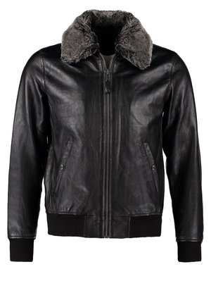 Homme portant une veste en cuir noire avec col en fourrure, t-shirt blanc à col en V, pantalon noir, bottes noires, portant un sac de voyage beige avec poignées marron.