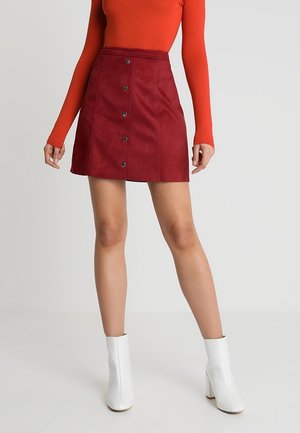 Femme portant une jupe en daim rouge boutonnée devant, un haut à manches longues orange et des bottines blanches, debout devant un fond uni.
