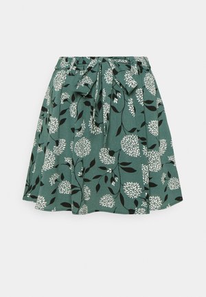 Jupe évasée verte avec motif floral blanc et ceinture nouée à la taille, présentant des détails de feuilles noires sur le tissu.