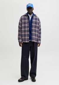 Nerti languotas marškiniai tamsiai mėlynos, raudonos ir kremų spalvų, dėvimi ant tamsiai mėlyno sportinio megztinio, derinami su plačiais tamsiais džinsais ir juodais batais. Mėlyna skrybėlė.