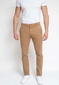 Hombre vestido con pantalones beige ajustados, camiseta blanca y zapatillas blancas, de pie contra un fondo blanco liso.
