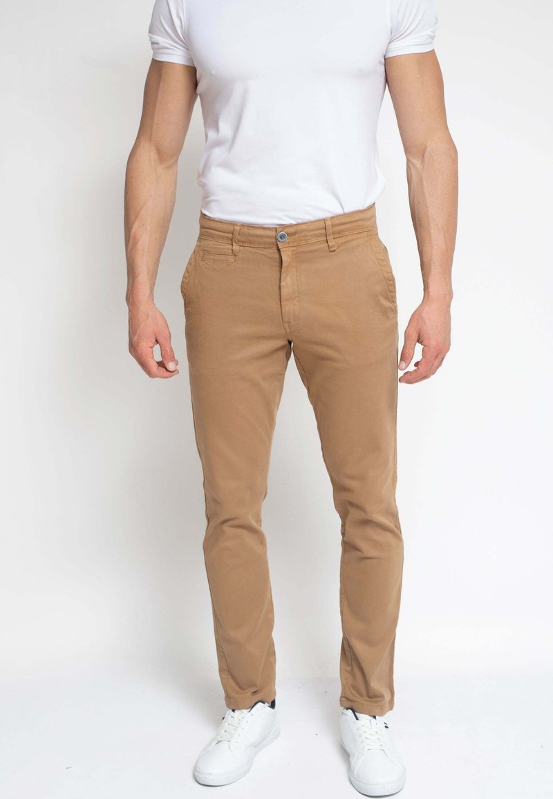 Hombre vestido con pantalones beige ajustados, camiseta blanca y zapatillas blancas, de pie contra un fondo blanco liso.