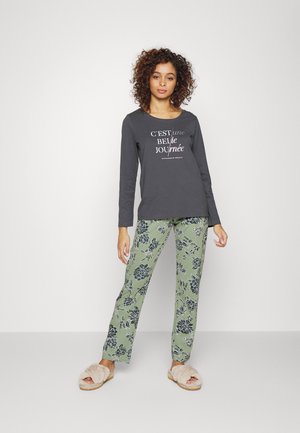 Langärmliges graues Oberteil mit einem weißen Schriftzug, kombiniert mit grünen Hosen mit floralen Mustern. Das Modell trägt helle Pantoffeln.