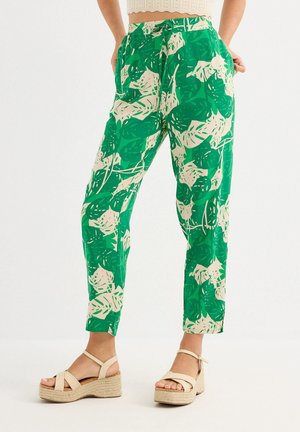 Mujer con pantalones de cintura alta con estampado tropical de hojas en verde y beige, y sandalias de plataforma beige, de pie contra un fondo blanco.