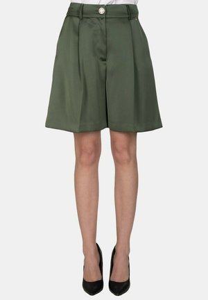 Persona che indossa shorts plissettati verde oliva al ginocchio, top bianco infilato dentro e décolleté nere a punta, in piedi su uno sfondo neutro.