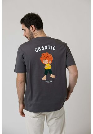Mann mit dunklem Haar, der ein dunkelgraues T-Shirt trägt, auf dem eine cartoonhafte rothaarige, barfüßige Figur mit verschränkten Armen abgebildet ist, darüber das Wort "GRANTIG".