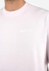 Rosa T-Shirt mit kurzen Ärmeln aus weichem Baumwollstoff, mit einem kleinen weißen "Pegador Co."-Logo auf der Brust. Angenehme Textur und klassischer Schnitt.