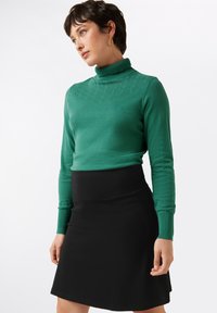 Groene turtleneck met een gestructureerd patroon bij de halslijn, gecombineerd met een nauwsluitende zwarte rok, beide gemaakt van gladde, zachte stoffen.
