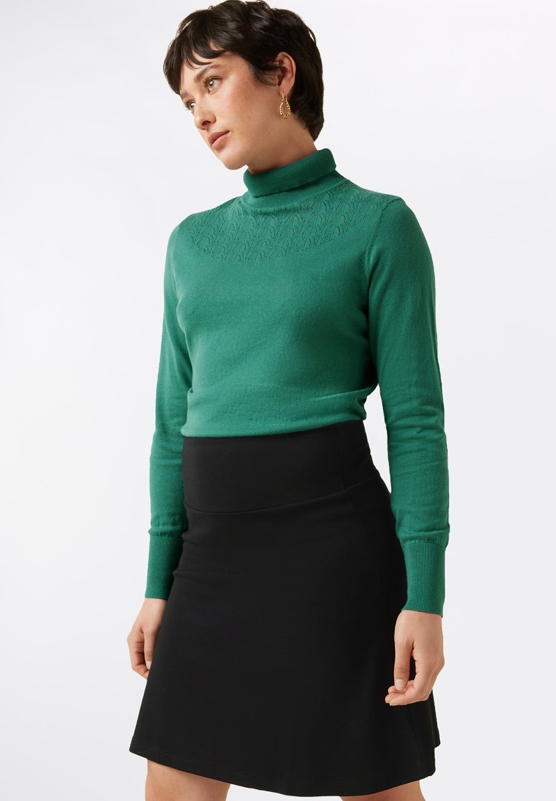 Groene turtleneck met een gestructureerd patroon bij de halslijn, gecombineerd met een nauwsluitende zwarte rok, beide gemaakt van gladde, zachte stoffen.