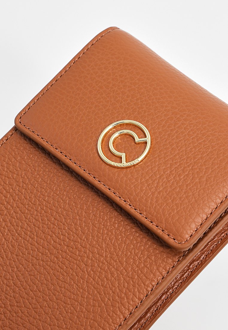 Brązowa torebka ze skóry o teksturowanej powierzchni, z okrągłym złotym logo i zapięciem na klapkę. Powierzchnia ma delikatne, wypukłe wzory oraz detale haftu.