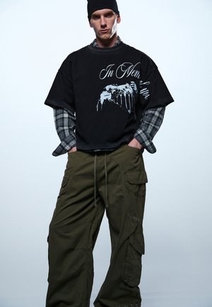 Bărbat purtând o căciulă neagră, un tricou negru stratificat cu imprimeu „In Memoriam” peste o cămașă gri în carouri și pantaloni lejeri cargo de culoare măslinie, stând cu mâinile în buzunare.