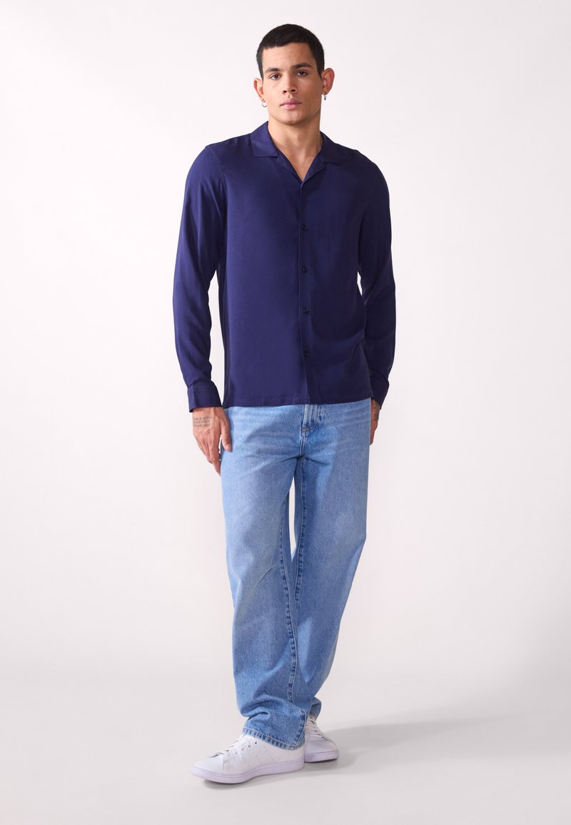 Marineblaues Hemd mit langen Ärmeln und Kragen, kombiniert mit hellblauen Jeans und weißen Turnschuhen. Glatte Textur, lässige Passform.