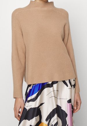 Femme portant un pull beige en maille côtelée à col montant et une jupe aux motifs abstraits colorés en bleu, noir, blanc et rose.