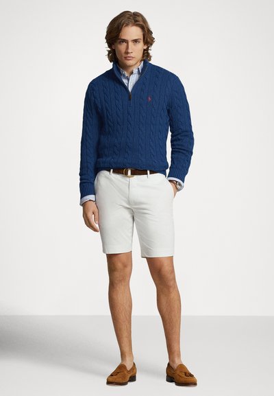 Μπλε πουλόβερ με πλέξη καλτσοδέτας, λευκά σορτς, καφέ ζώνη και καφέ μοκασίνια. Πουκάμισο με μπλε και λευκές ρίγες ορατό στο γιακά και τα μανίκια.