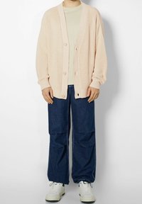 Cardigan en tricot rose clair avec texture côtelée, coupe oversize et deux boutons, associé à un jean large bleu et des baskets blanches.