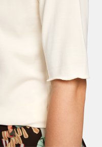 Cream short-sleeve top made from soft fabric, featuring a scalloped edge on the sleeves. Light texture and fitted design.