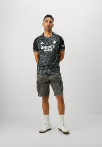 Svart idrettsjersey med grafisk mønster, hvit tekst og grå cargo shorts. Bært med hvite joggesko og ribbede sokker.