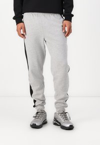 Pantaloni de trening gri cu dungi negre pe laterale, talie elastică și picioare conice, asortați cu pantofi sport argintii. Material dintr-un amestec de bumbac fin.