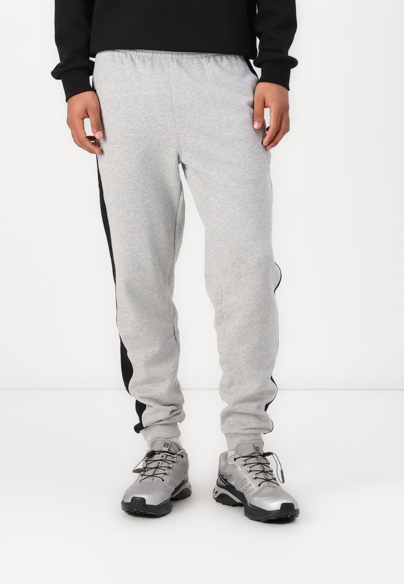 Pantaloni de trening gri cu dungi negre pe laterale, talie elastică și picioare conice, asortați cu pantofi sport argintii. Material dintr-un amestec de bumbac fin.