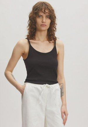 Schwarzes Baumwoll-Top mit dünnen Trägern. Kombiniert mit weißen Jogginghosen mit Kordelzug. Das Model steht mit den Händen in den Taschen, der Stoff hat eine strukturierte Textur.