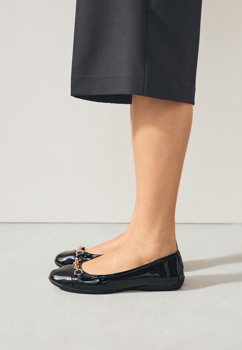 Femme portant des ballerines en cuir verni noir avec un détail chaîne dorée et un pantalon noir raccourci, debout sur un sol clair contre un mur uni.