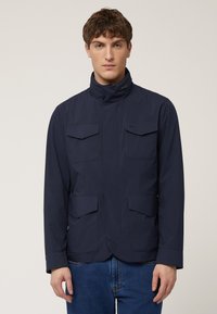 Navyblauwe jas met een hoge kraag, vier voorzakken en een gladde textuur. Het ontwerp heeft een ritssluiting aan de voorkant en lange mouwen.