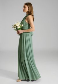 Robe longue et fluide en tissu vert doux avec un col halter torsadé et des détails en dentelle. Le mannequin tient un bouquet de fleurs blanches.