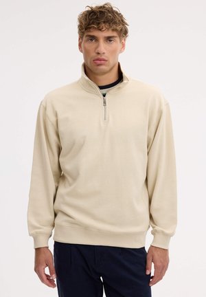 Un hombre luce un jersey color crema con cremallera de un cuarto y cuello elevado, junto con unos pantalones oscuros, sobre un fondo blanco liso.