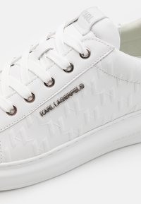 Hvit sneakers med teksturert syntetisk overdel som har et geometrisk mønster, sølvfarget merkevare og kontrasterende hvite lisser og såle.