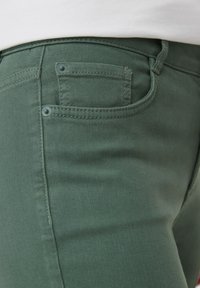 Zielone dżinsy skinny w gładkiej teksturze, z małą tylnią kieszenią i detalem w postaci przeszycia, zabezpieczone jasnoniebieskimi akcesoriami.