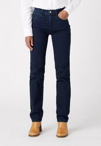 Donkerblauwe spijkerbroek met een slim fit, hoge taille, voorzakken en stikselaccenten, gecombineerd met tan leren laarzen met hak.