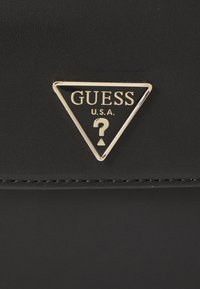 Faux leather nero con superficie texturizzata, presenta un logo triangolare dorato con "GUESS" e un punto interrogativo al centro.