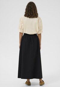 Boleró barna göndör hajú nő, bézs rövid ujjú blúzt, fekete hosszú szoknyát és barna slip-on cipőt viselve, háttal áll.