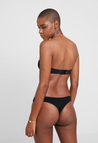 Vrouw met kort haar in een zwart strapless bikini, van achteren gezien tegen een effen lichte achtergrond.
