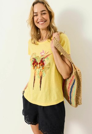 Smiling woman wearing yellow shirt with colorful tropical birds, black shorts with eyelet hem, and carrying woven tote bag with rainbow stripes.