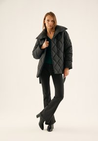 Black quilted puffer jacket with a high collar and snap closures, paired with a green top and flared black jeans, worn with heeled black boots.