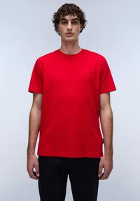 Maglietta rossa in cotone con maniche corte, scollo rotondo e piccolo logo in rilievo sul lato sinistro del petto. Pantaloni neri, sfondo semplice.