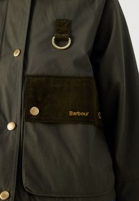 Olijfgroene waterdichte jas met gouden knopen en een corduroy zak. Bevat een metalen ring en "Barbour" geborduurd in goud.