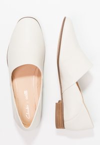 Chaussures slip-on en cuir blanc avec un design courbé, un petit talon empilé, et une finition lisse. Equipées d'une semelle intérieure matelassée pour le confort.
