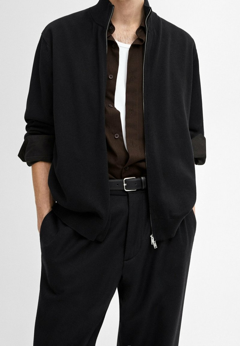Homme portant une veste noire zippée par-dessus une chemise marron foncé déboutonnée et un sous-vêtement blanc, avec un pantalon noir et une ceinture, les mains dans les poches.