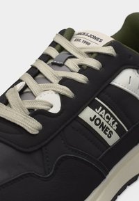 Μαύρο και άσπρο sneaker Jack & Jones με μπεζ κορδόνια, ετικέτα με την επωνυμία στη πλευρά και πράσινη εσωτερική επένδυση ορατή κοντά στη γλώσσα.