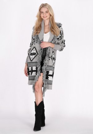 Cardigan grigio lavorato a maglia con motivi geometrici neri, bordi frangiati e colletto a scialle, indossato sopra una maglietta bianca e abbinato a stivali neri.