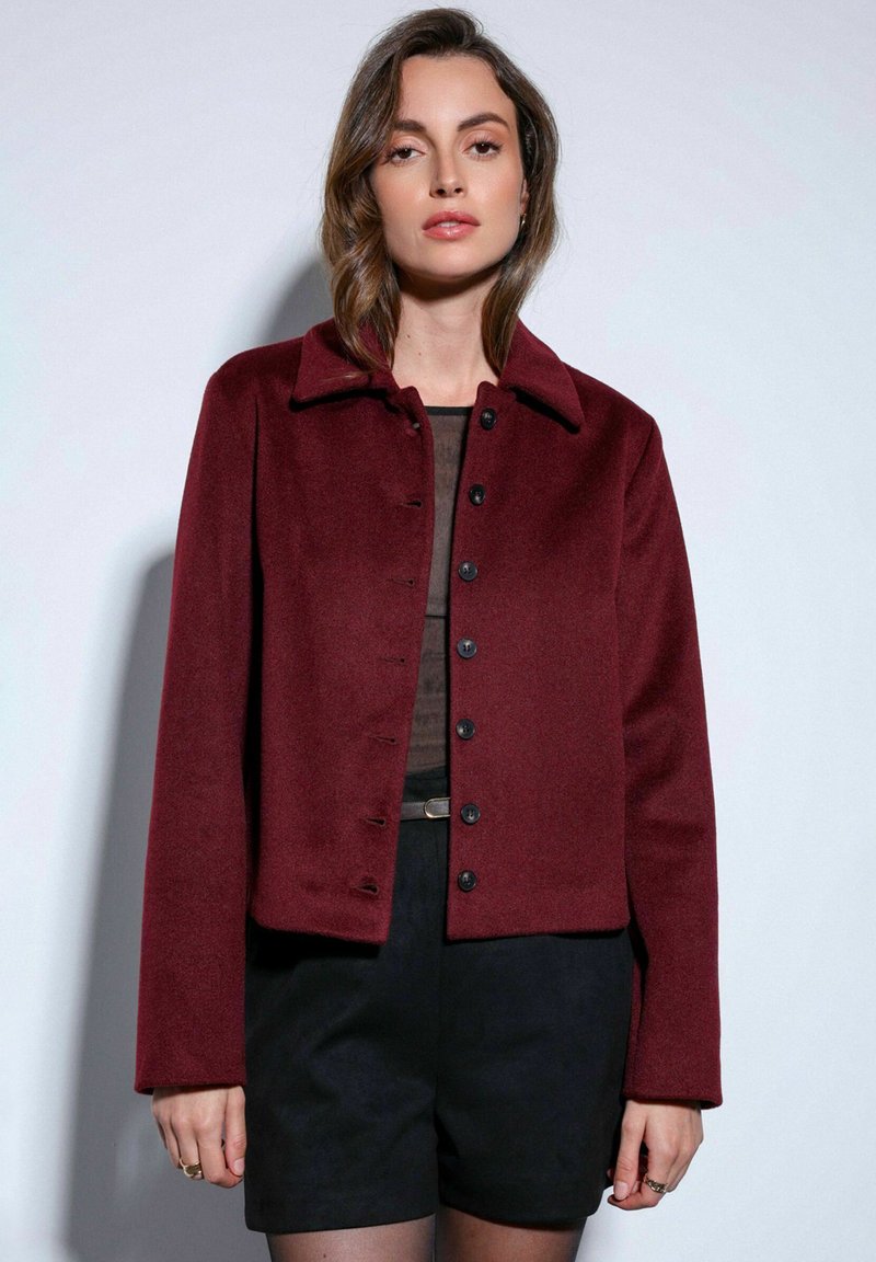 Bordeauxrode cropped jas met een kraag, knoopsluiting aan de voorkant en een zachte textuur, gecombineerd met zwarte shorts en een doorschijnende top.