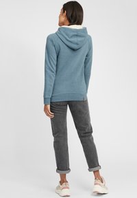 Sudadera azul con un forro interior color crema, que presenta una capucha con cordón, mangas largas y una pequeña etiqueta marrón en el dobladillo. Combinada con pantalones gris con puños doblados.