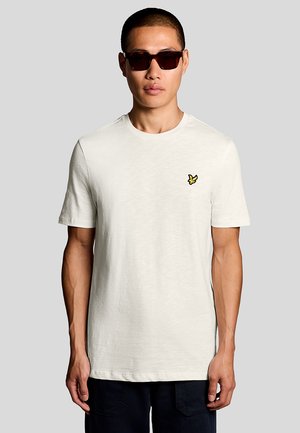 Mand iført en enkel hvid t-shirt med en lille gul fuglelogo på brystet og mørke solbriller, stående mod en neutral baggrund.
