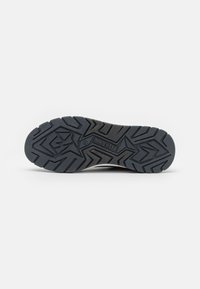 Black rubber outsole with textured tread pattern featuring grooves and a logo. Designed for traction and stability in various conditions.