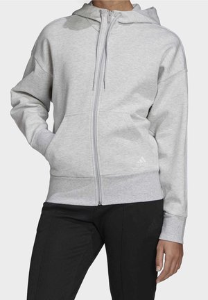 Persoon draagt lichtgrijs sweatvest met volledige ritssluiting en capuchon, voorzakken en zwarte broek, staand tegen een effen achtergrond.