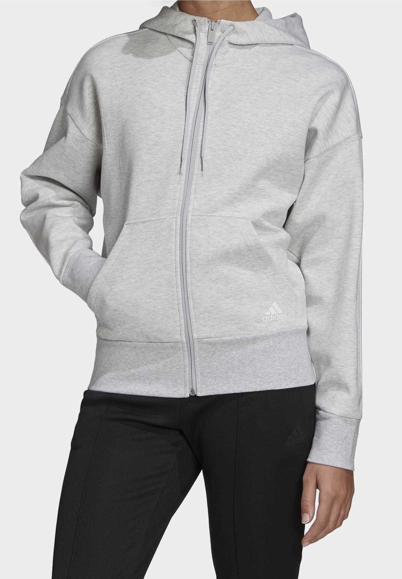 Personne portant un sweat à capuche gris clair avec fermeture éclair intégrale et poches avant, et un pantalon noir, debout devant un fond uni.